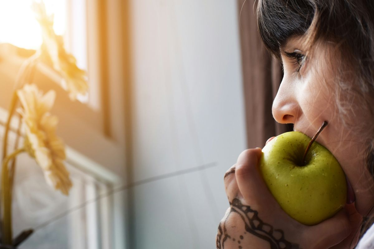 A profile view of a person about to bite into a green apple, with sunlight highlighting their face and a visible tattoo on their hand. Flowers are in soft focus in the foreground.